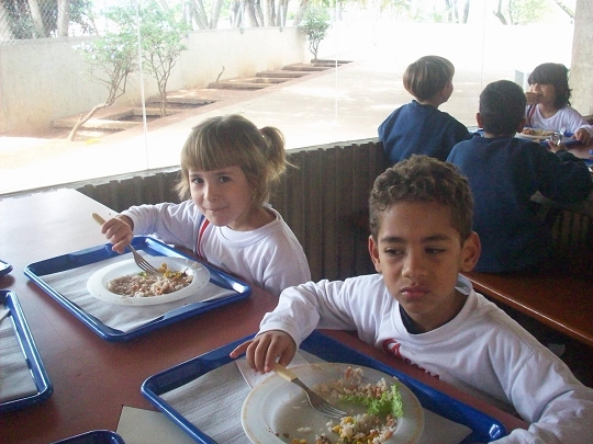 Crianças almoçando no refeitório com bandejas, na reportagem sobre alimentação