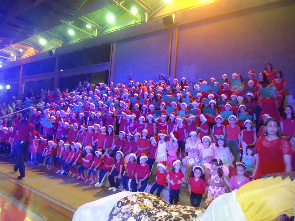 Apresentação da cantata O Auto do Menino Redentor no ginásio, com alunos de vermelho e branco sob luz cênica