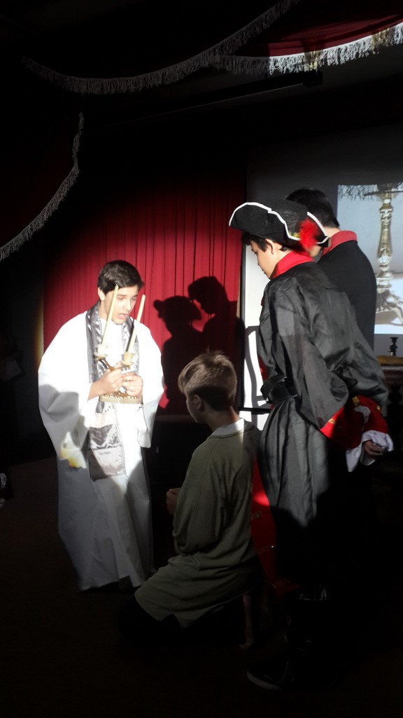 Alunos em cena de teatro com capa vermelha sob a luz do palco