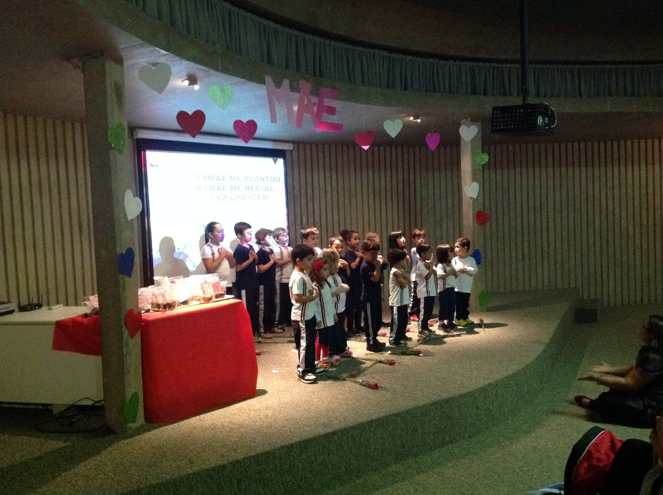 Alunos do Período Integral enfileirados no palco, diante do painel decorado com a palavra Mãe, na apresentação do Dia das Mães