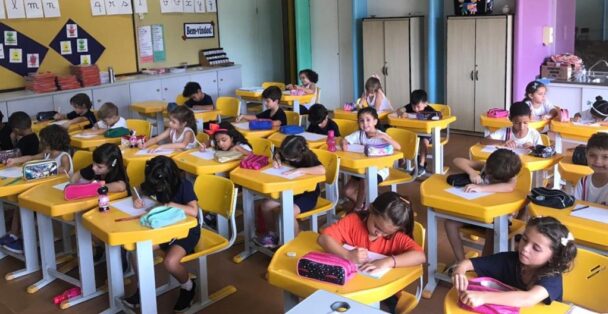Sala de aula do fundamental com alunos em carteiras amarelas durante atividade de alfabetização
