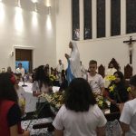 Alunos vestidos de branco em procissão pelo corredor da igreja, na Coroação de Nossa Senhora