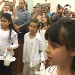Alunos vestidos de branco em procissão pelo corredor da igreja, na Coroação de Nossa Senhora