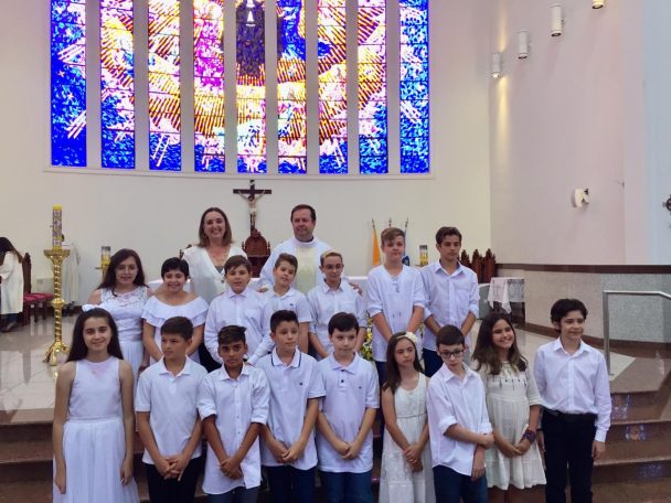 Turma de branco posando na capela diante dos vitrais na celebração da Primeira Eucaristia