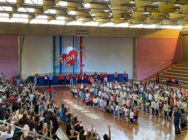 Ginásio cheio de alunos na homenagem do Dia das Mães, com um coração projetado no telão