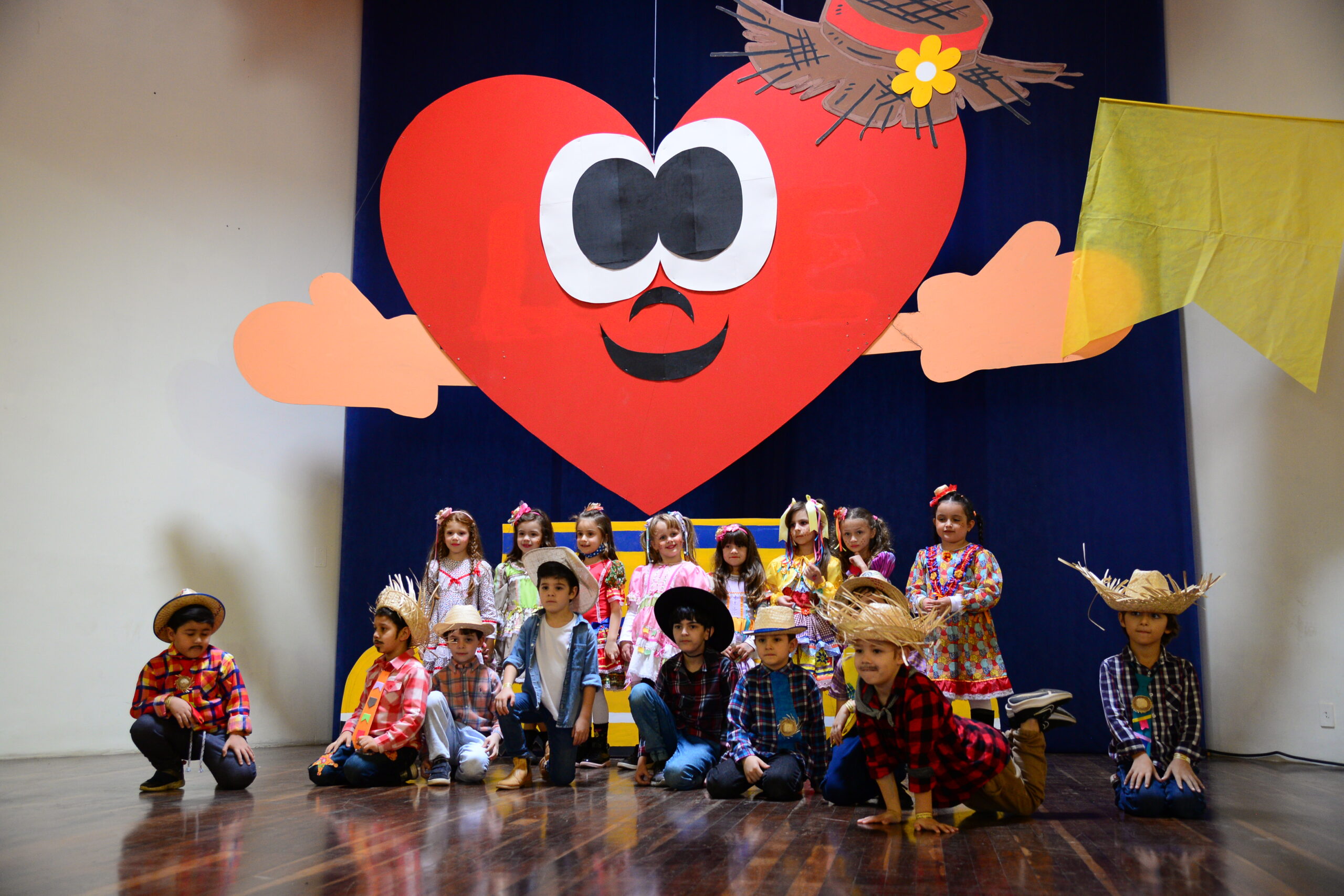 Apresentação da educação infantil no palco do Arraiá do Coração, com cenário de coração gigante e trajes juninos