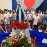 Alunos em volta da imagem de Nossa Senhora enfeitada com flores vermelhas na celebração do mês de Maria