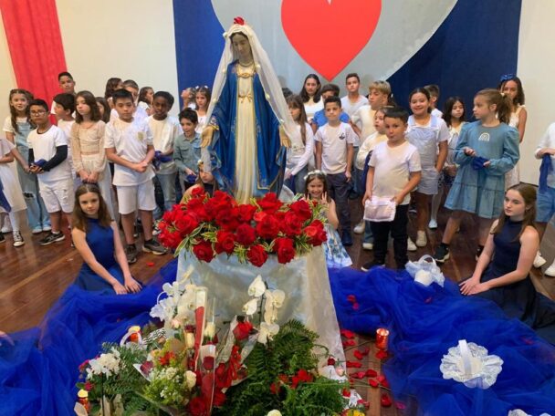 Alunos em volta da imagem de Nossa Senhora enfeitada com flores vermelhas na celebração do mês de Maria
