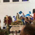 Alunos erguem cartazes azuis diante do altar florido na celebração do Mês de Maria