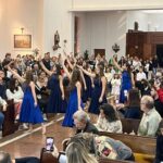 Alunas de vestido azul cantando na capela lotada durante a celebração do mês de Maria