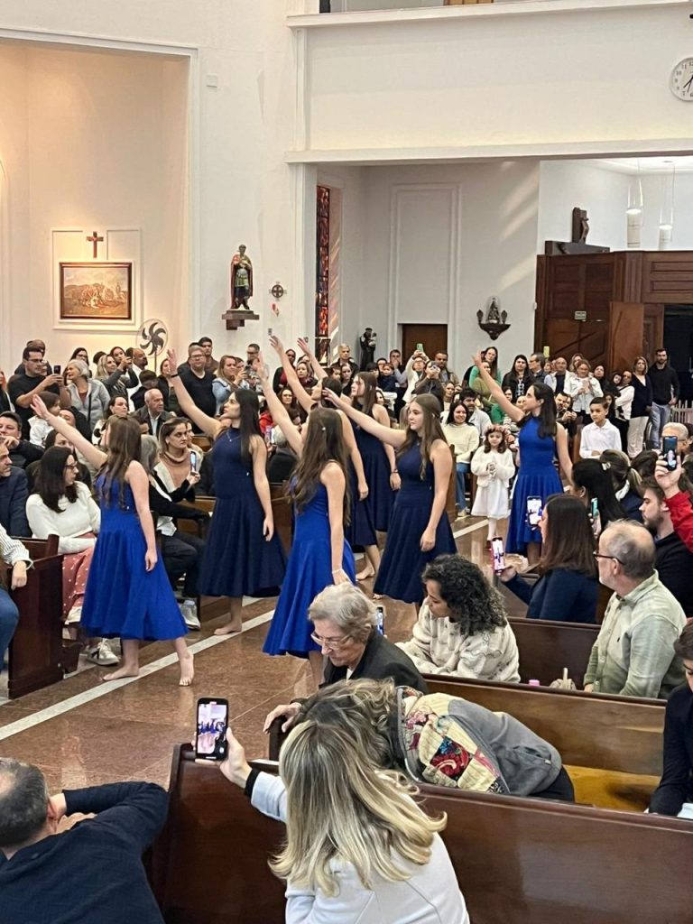 Alunas de vestido azul cantando na capela lotada durante a celebração do mês de Maria
