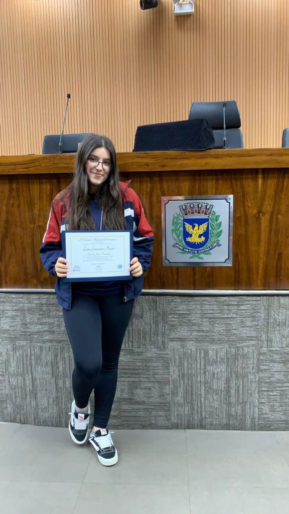 Aluna segurando o certificado do projeto Parlamento Jovem no plenário da Câmara Municipal