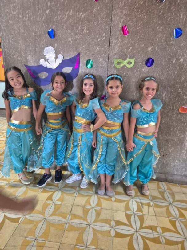 Cinco meninas fantasiadas com traje azul diante do painel de máscaras no Carnaval do Coração