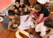 Alunos e professora decorando no chão da sala a camiseta branca da homenagem Mamãe Camisa 10