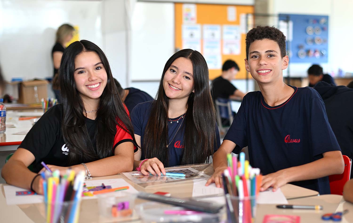 Três estudantes de uniforme sorrindo lado a lado na mesa de trabalho da sala de aula, com potes de canetas coloridas e material espalhado à frente