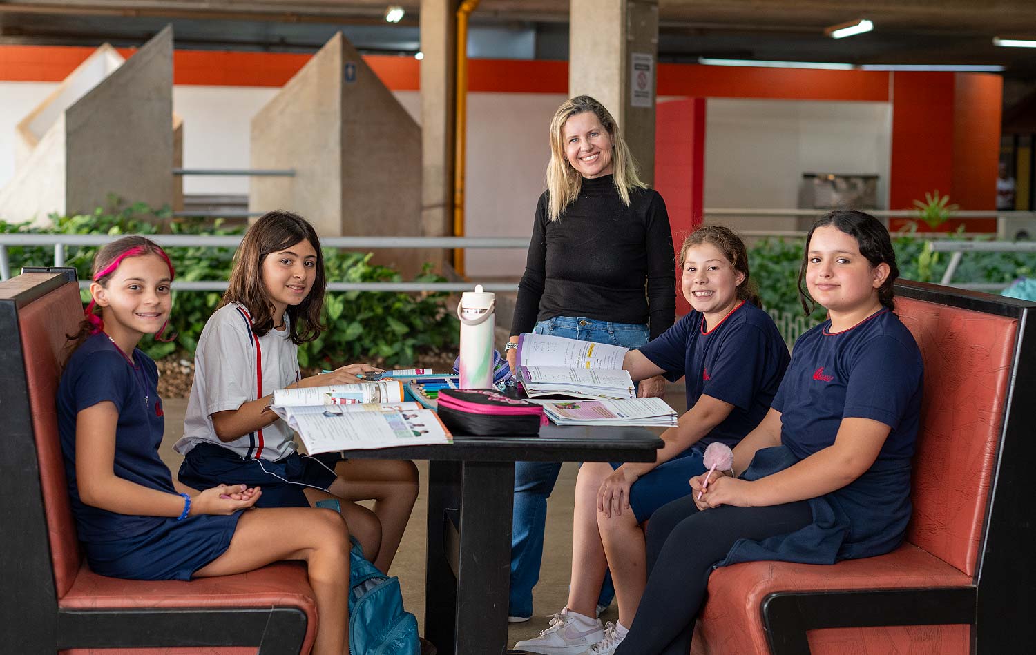 Quatro estudantes de uniforme sentadas em poltronas ao redor de uma mesa de estudo, com livros e cadernos abertos, e uma professora em pé ao fundo, na área de convivência coberta do colégio