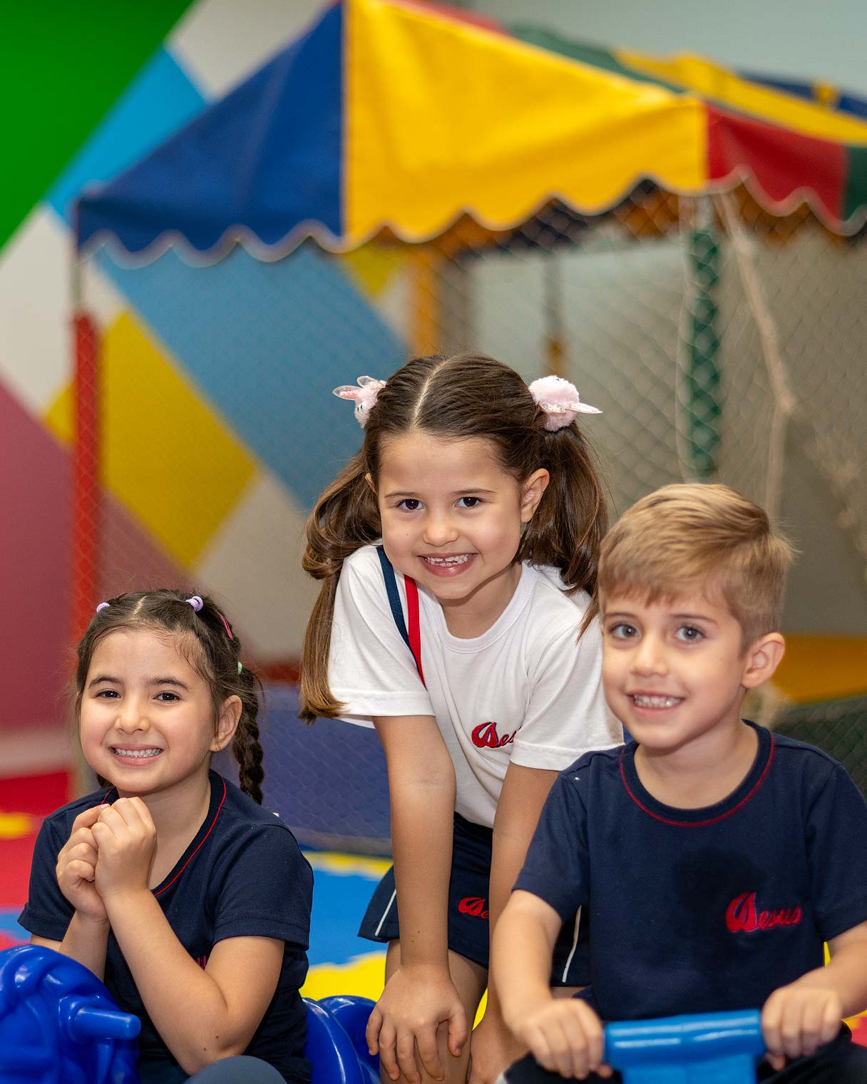 Três crianças da Educação Infantil sorrindo na brinquedoteca colorida, sentadas em brinquedos de montar, com tenda listrada e rede de escalada ao fundo