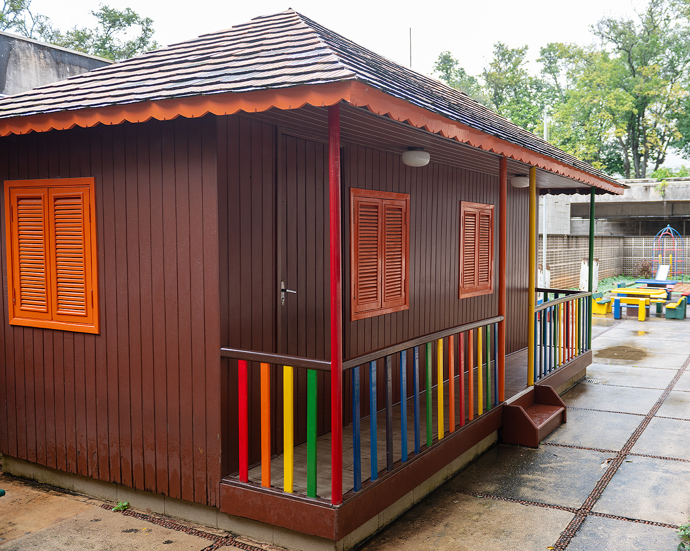 Casinha de madeira da Educação Infantil, pintada de marrom com janelas laranja e gradil colorido na varanda, cercada por árvores