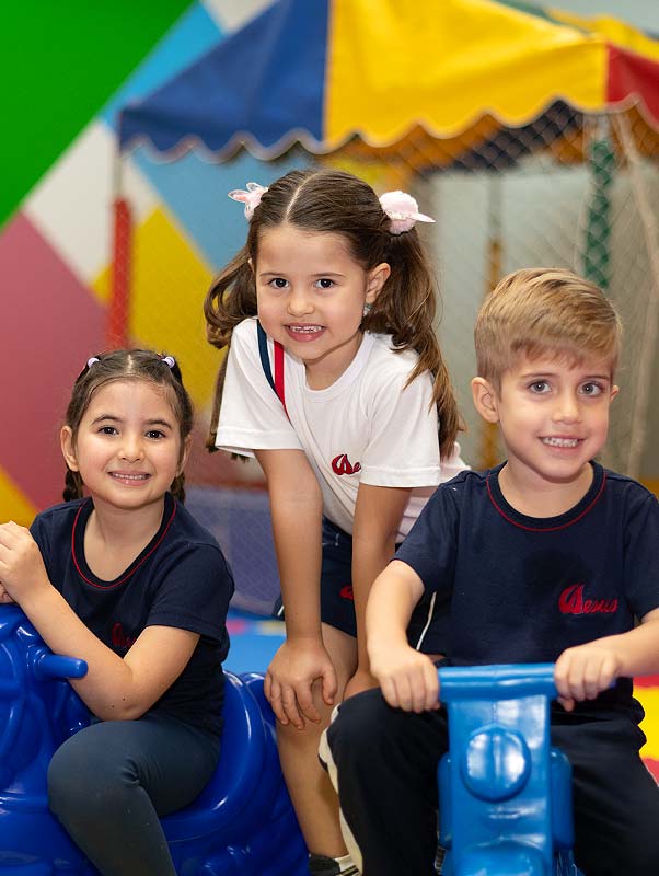 Três crianças da Educação Infantil de uniforme sorrindo sobre brinquedos azuis, com a tenda colorida do espaço ao fundo