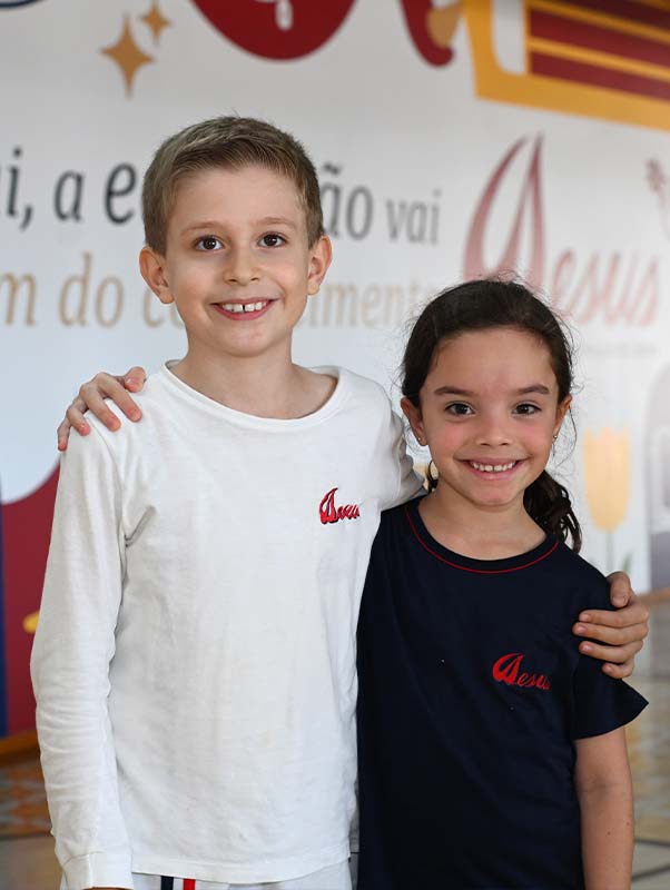 Dois alunos do Ensino Fundamental I de uniforme, abraçados e sorrindo, diante do painel da campanha do colégio