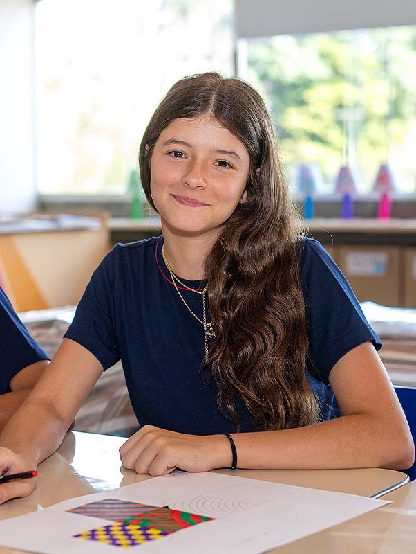Estudante do Ensino Fundamental II de uniforme azul sorrindo sentada na carteira, com um desenho colorido de formas geométricas sobre a folha à sua frente