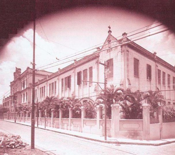 Fotografia antiga em sépia da fachada original do colégio, prédio de dois andares na esquina, com palmeiras atrás do muro, poste e fios sobre a rua ainda de paralelepípedo