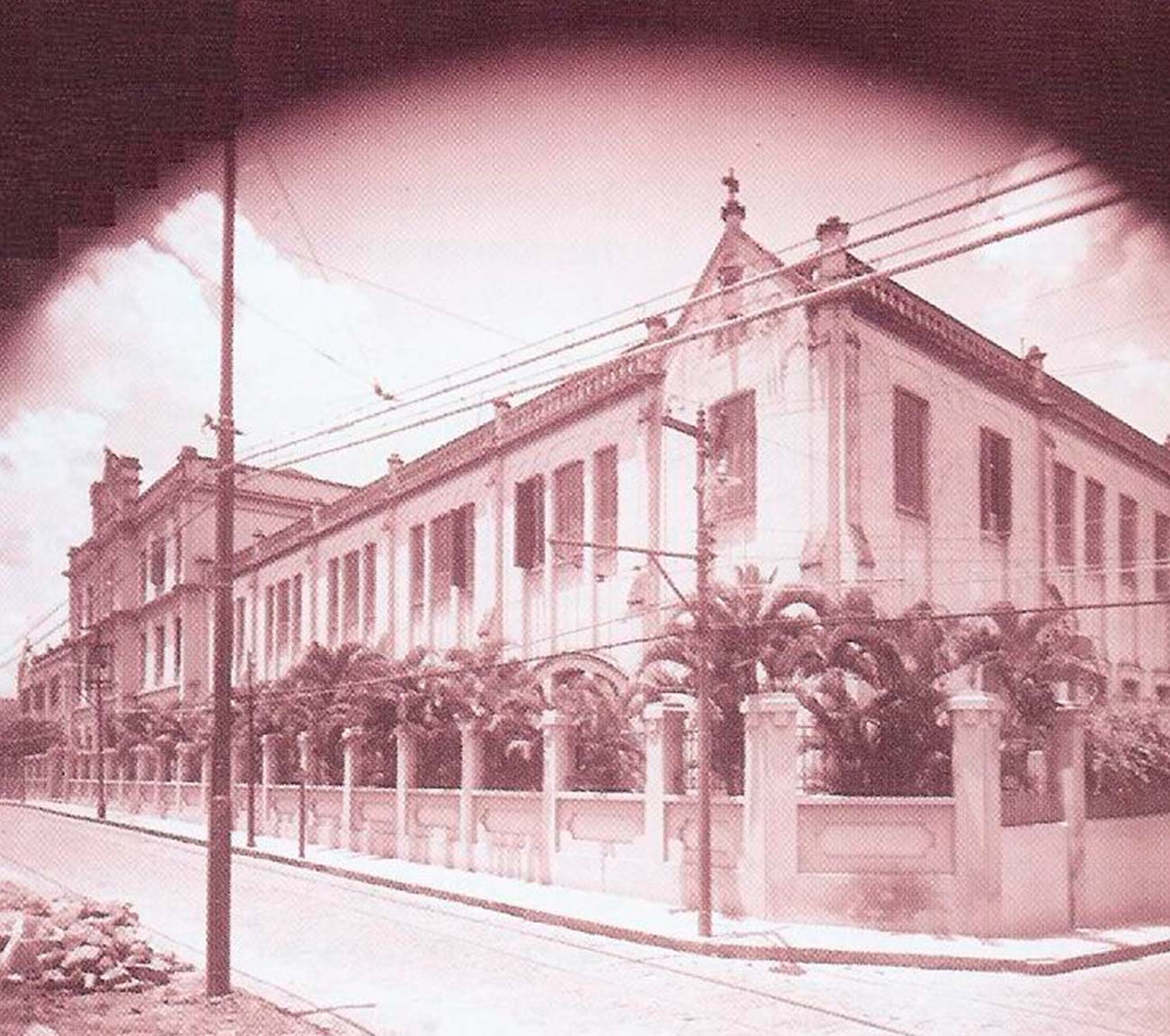 Fotografia antiga em sépia da fachada original do colégio, prédio de dois andares na esquina, com palmeiras atrás do muro, poste e fios sobre a rua ainda de paralelepípedo