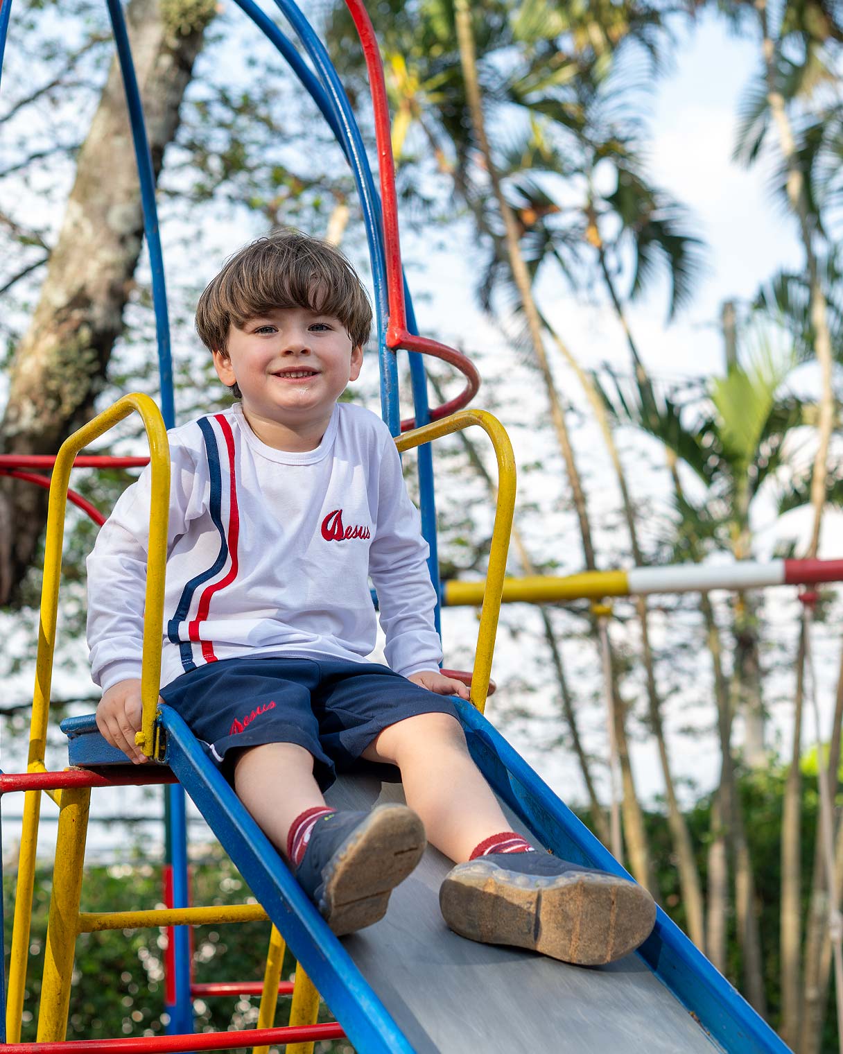 Menino de uniforme branco e azul sorrindo sentado no alto do escorregador do parque, entre as barras coloridas e as árvores ao fundo