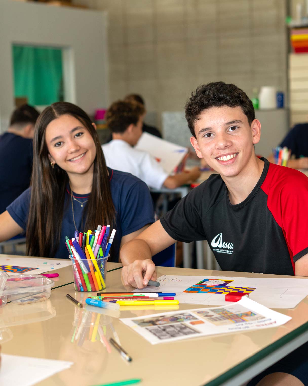 Aluna de uniforme azul e aluno de camiseta preta e vermelha do Ensino Fundamental II sorrindo em uma mesa de sala de aula, com canetinhas coloridas e trabalhos sobre a mesa