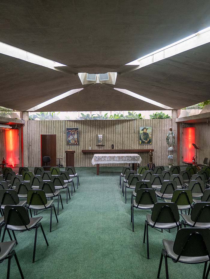 Capela do colégio com fileiras de cadeiras voltadas para o altar, teto em concreto e luz natural entrando pelo fundo