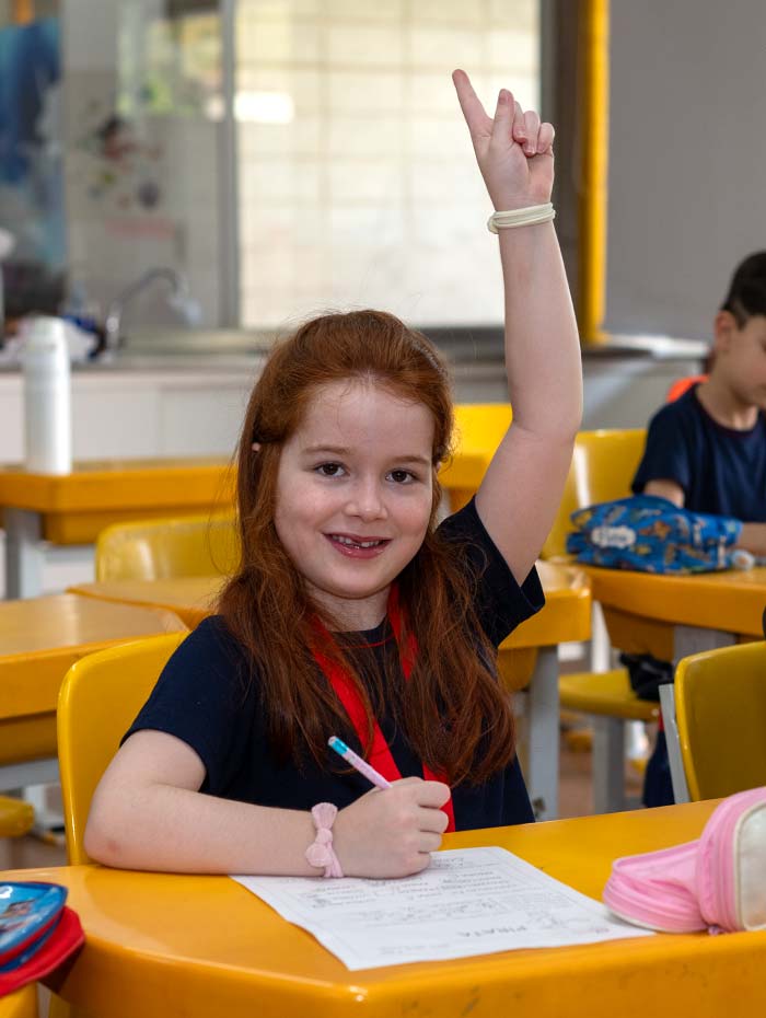 Aluna ruiva de uniforme sorrindo com a mão levantada na sala de aula, caderno e caneta sobre a mesa amarela