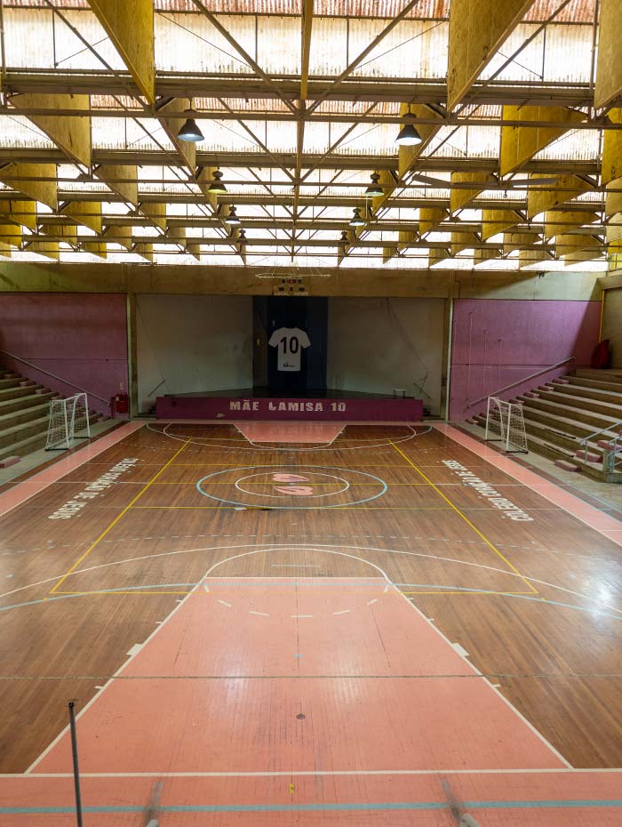 Ginásio poliesportivo coberto do colégio, com quadra demarcada, arquibancada e estrutura metálica amarela no teto