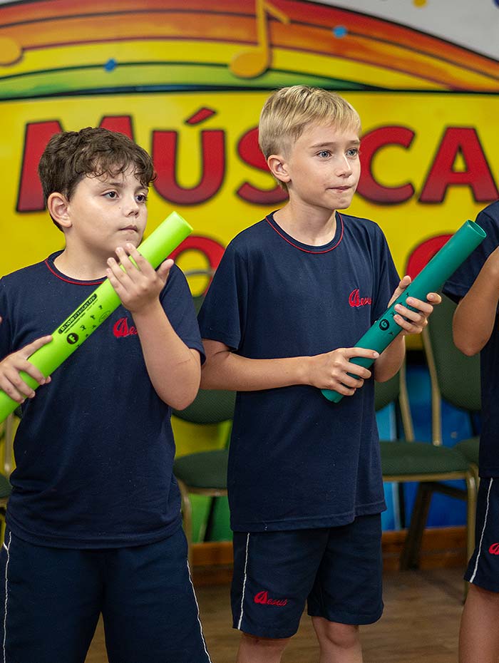 Dois alunos de uniforme tocando flauta na oficina de música, diante do painel amarelo com notas musicais