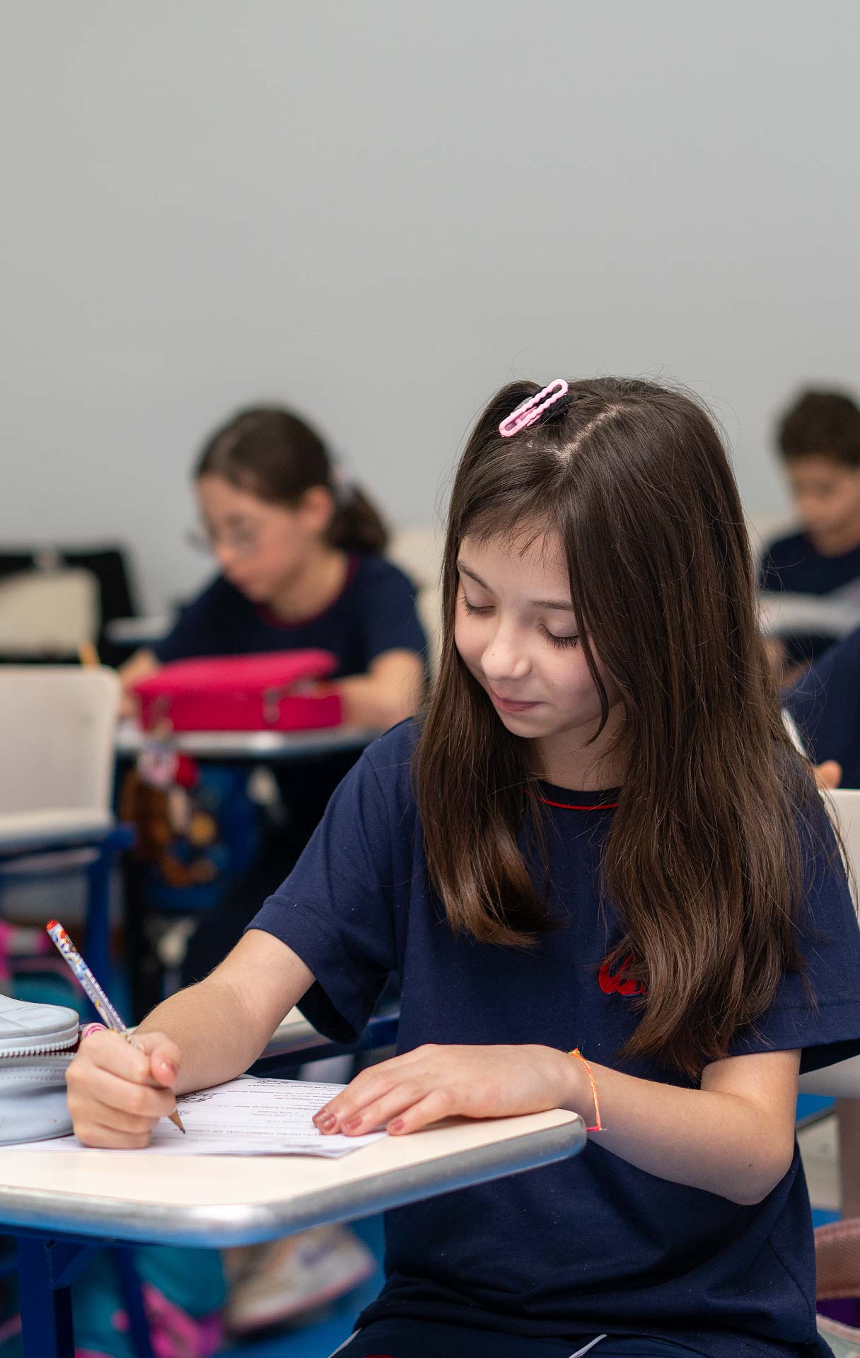 Aluna de uniforme azul escrevendo com lápis na carteira durante a aula, com outros colegas estudando ao fundo