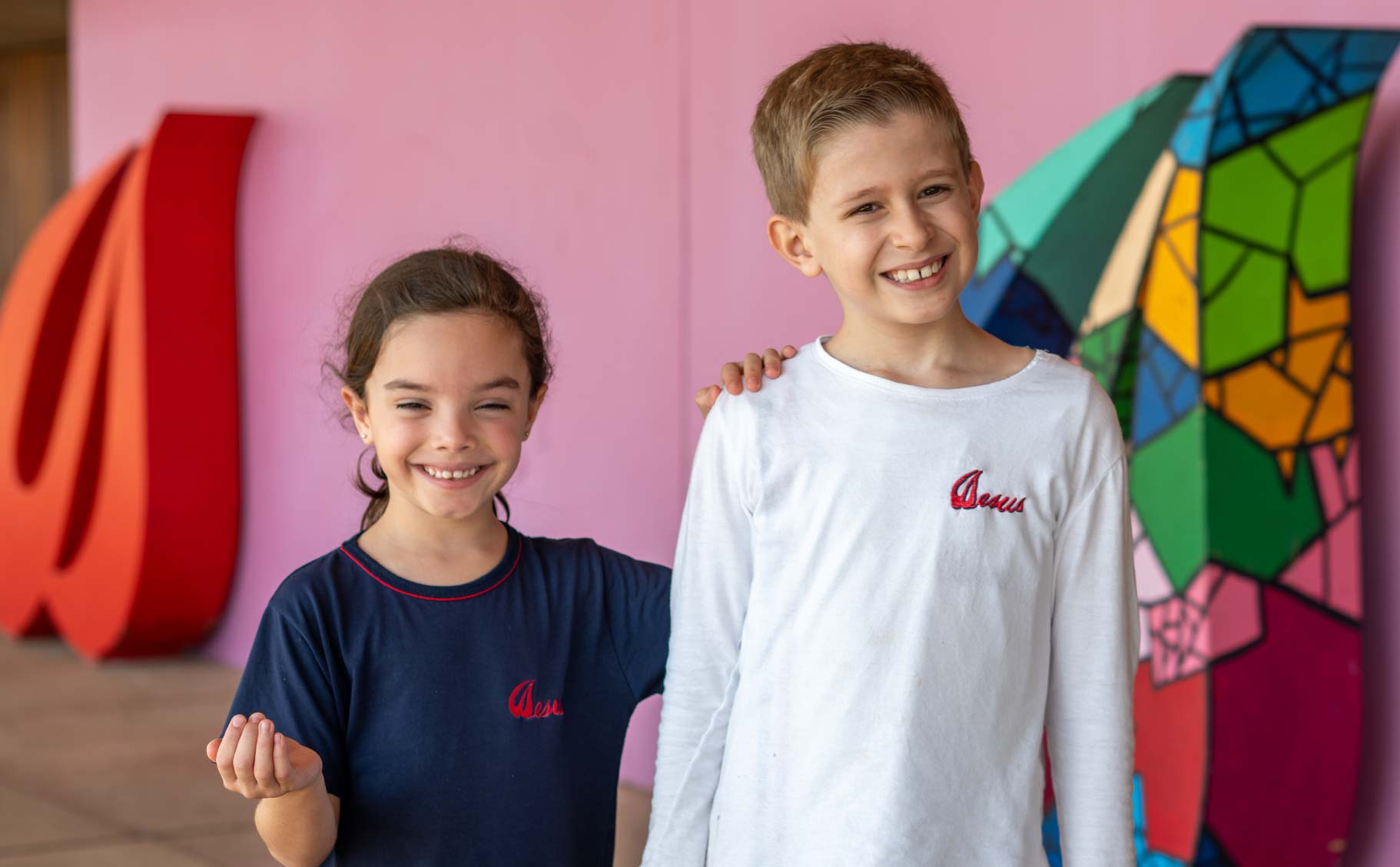 Menina de uniforme azul e menino de uniforme branco do Ensino Fundamental I sorrindo diante de uma parede rosa com escultura colorida