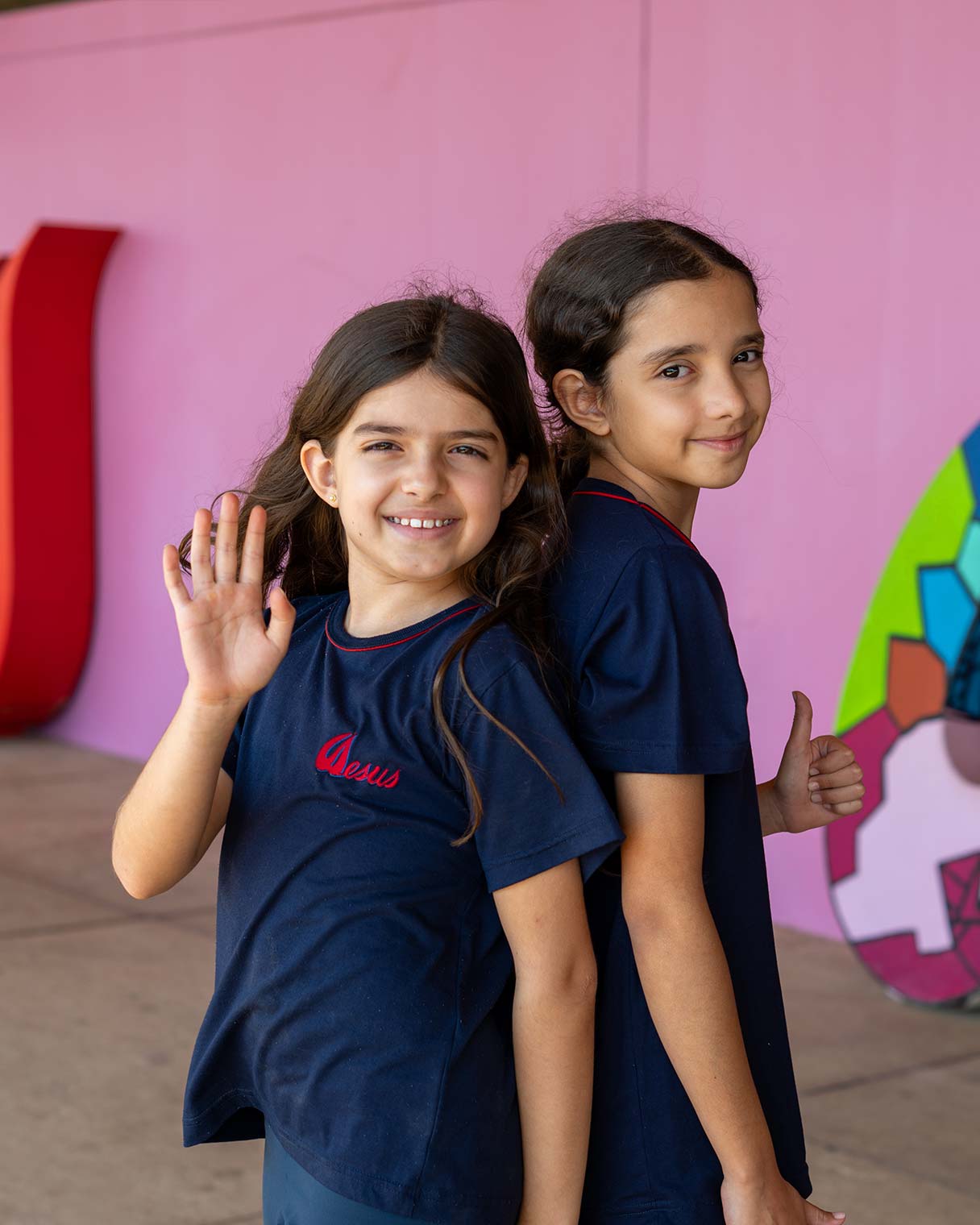 Duas meninas do Ensino Fundamental I de uniforme azul, de costas uma para a outra, uma acenando e a outra fazendo sinal de positivo, diante de um painel rosa com mural colorido