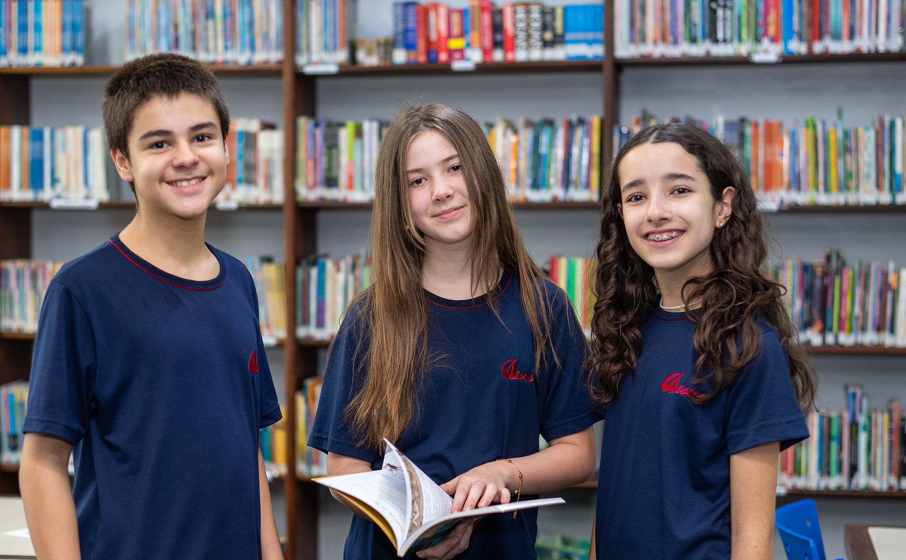 Três alunos do Ensino Fundamental II de uniforme azul sorrindo na biblioteca, a do meio segurando um livro aberto, com estantes cheias de livros ao fundo