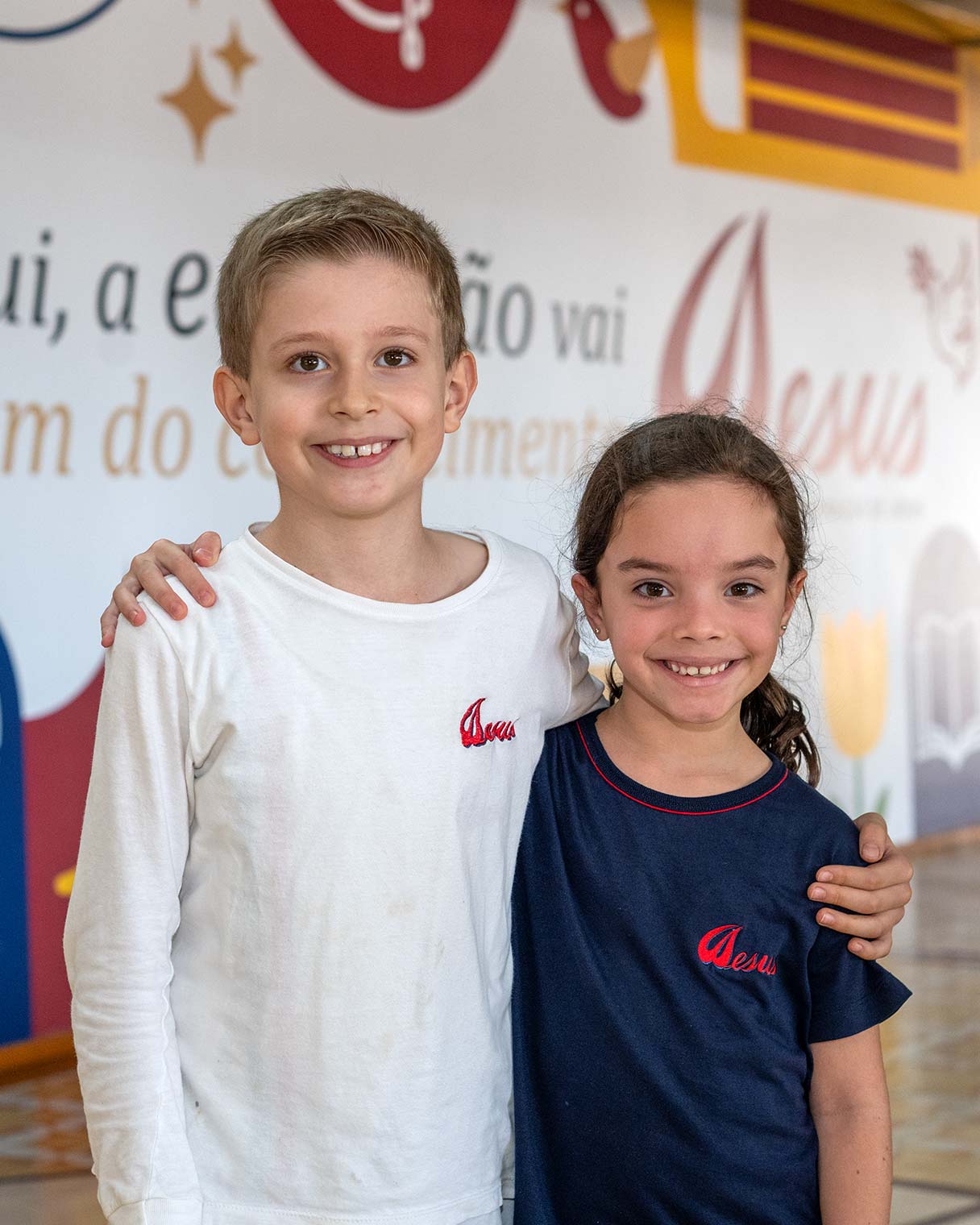 Menino de uniforme branco com o braço no ombro de uma menina de uniforme azul, ambos sorrindo diante de um painel pintado na parede da escola