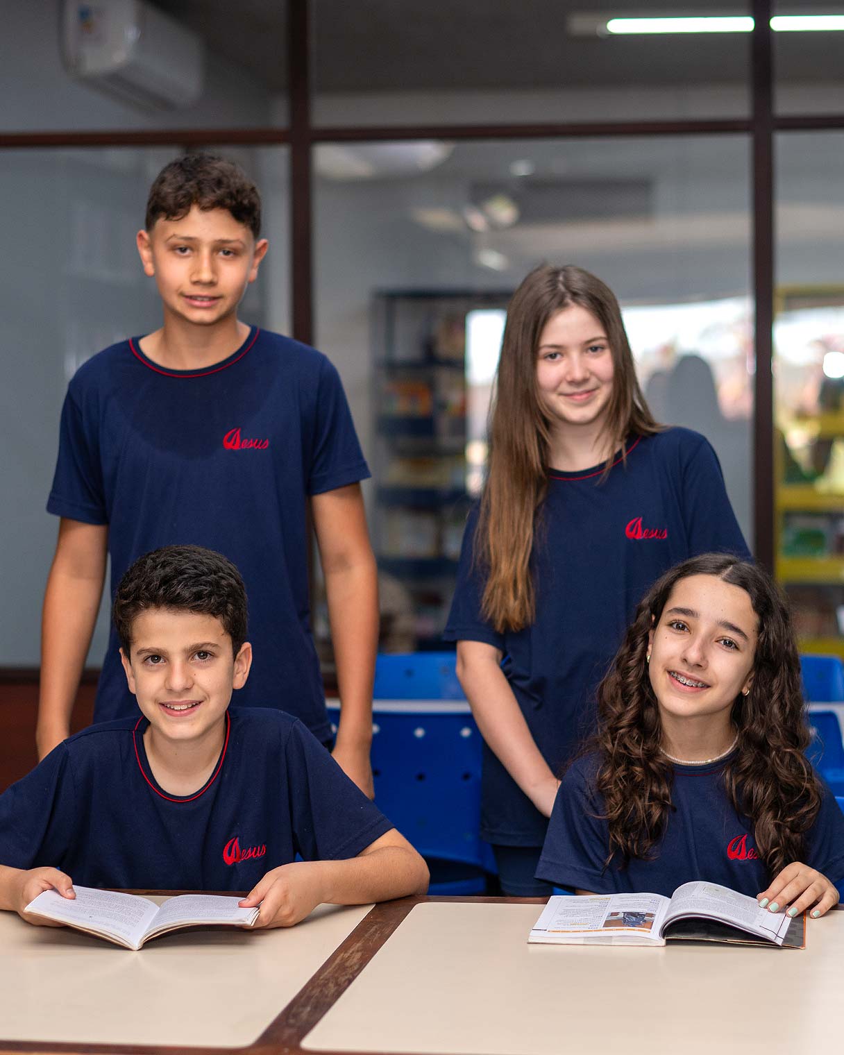 Quatro alunos do Ensino Fundamental II de uniforme azul sorrindo na sala de aula, dois sentados com livros abertos sobre a carteira e dois em pé atrás