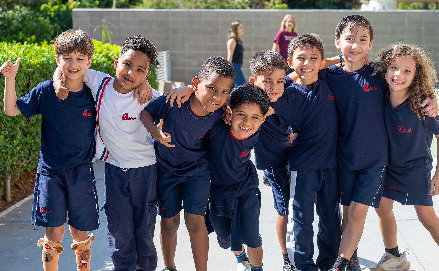 Grupo de crianças da Educação Infantil de uniforme azul, abraçadas e sorrindo em um caminho externo do colégio, com arbustos ao fundo