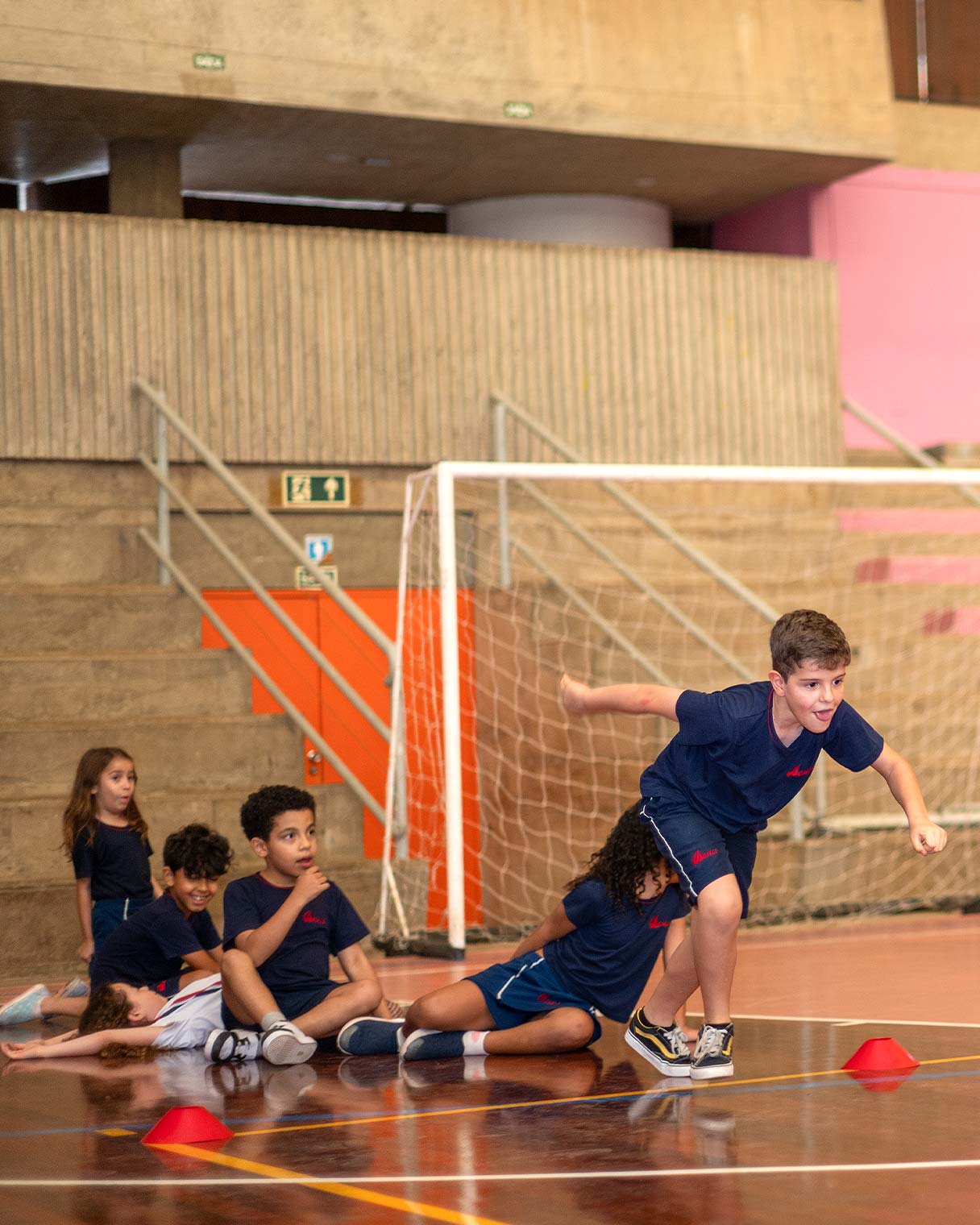Crianças do Período Integral de uniforme esportivo azul na quadra, um menino correndo em primeiro plano e os colegas sentados no chão, ao lado do gol e dos cones