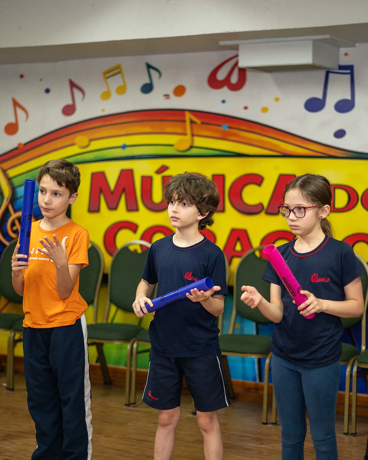 Três crianças do Período Integral em pé com tubos sonoros coloridos nas mãos, diante de um painel pintado com notas musicais e os dizeres Música do Coração