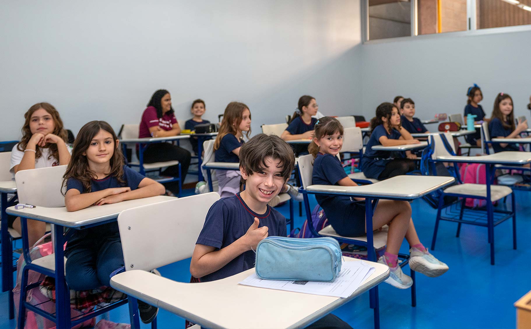 Sala de aula do Período Integral com alunos de uniforme azul em carteiras individuais, um menino em primeiro plano sorrindo e fazendo sinal de positivo, professora ao fundo