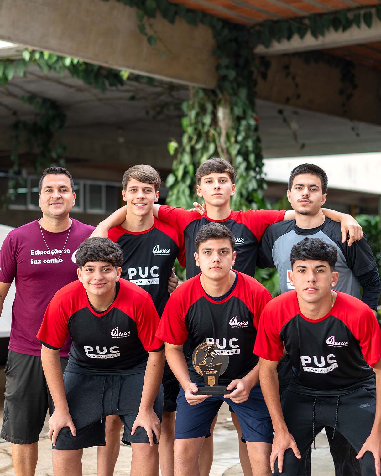 Equipe de futsal do Ensino Médio posando com o troféu ao lado do professor, todos de uniforme esportivo preto e vermelho, em uma área externa
